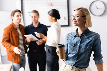 young, cheerful businesswoman in eyeglasses holding coffee to go while multicultural colleagues talking on blurred background