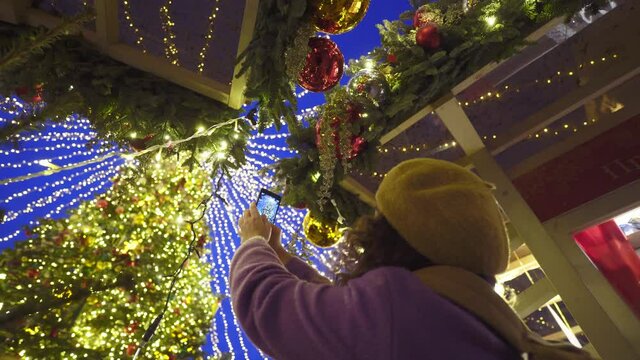 Young curly haired woman in beret takes picture of wonderful Christmas tree spending time in decorated amusement park at winter fair in evening low angle shot