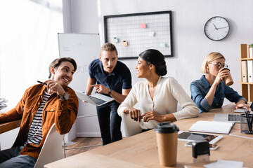 young manager pointing at laptop near multicultural colleagues working together in office, blurred foreground