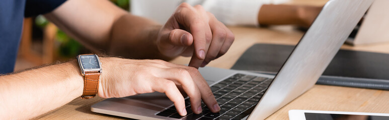 Cropped view of man with watch typing on laptop, while sitting at desk with blurred african american woman on background, banner