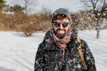 Fototapeta na wymiar Happy hipster man with beard and backpack having fun in winter park during a snowfall. Wearing sunglasses.