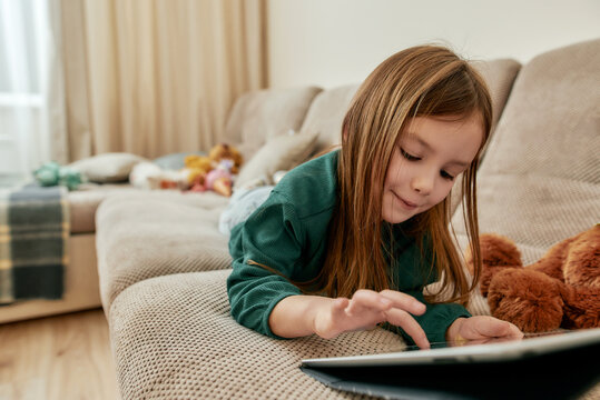 A cute little girl enjoy playing games on her tablet while lying on her stomach on a sofa