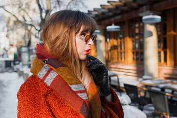 Close up winter portrait of elegant brunette woman in retro glasses posing outdoor in old European city. Standing near cafe. Wearing brown  coat, wool scarf and leather gloves.