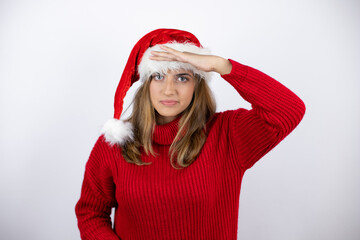 Young pretty blonde woman wearing a red casual sweater and a christmas hat over white background very happy and smiling looking far away with hand over head. searching concept.