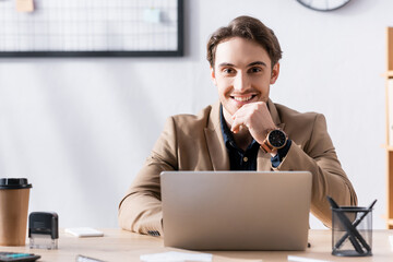 Smiling office worker looking at camera while sitting near laptop at desk in office on blurred background