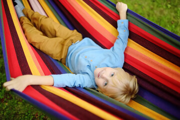 Fototapeta na wymiar Cute little blond caucasian boy relaxing and having fun in multicolored hammock in backyard or outdoor playground. Summer active leisure for kids. Child swinging on hammock.