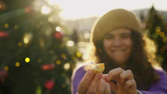 Positive young woman in purple coat and beret breaks fortune cookie and shows paper sheet against blurred Christmas trees in evening closeup