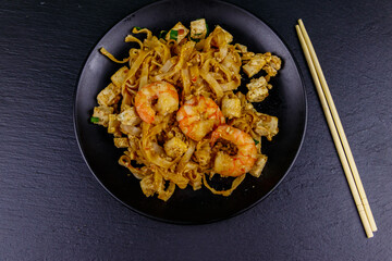 Stir-fried rice noodles Pad Thai with prawns and tofu in a plate on black slate background. Top view. Thai food