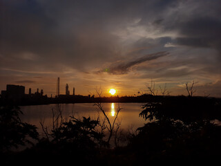 the sun sets over the lake and among the factories and trees