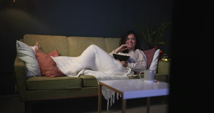 A modern young woman lies on the couch watching a movie and laughing. A girl with popcorn lies on the couch late at night watching TV shows. TV series, movies.