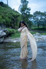 Fototapeta na wymiar Beautiful Indian young female model wearing traditional saree standing in front of a river landscape with overcast clouds.