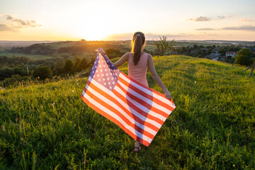 Happy young woman posing with USA national flag standing outdoors at sunset. Positive girl celebrating United States independence day. International day of democracy concept.