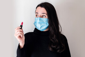 Una mujer joven morena lleva una mascarilla quirúrgica azul y sujeta un pintalabios rojo