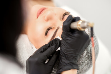Beautician applying permanent makeup on eyebrows of young woman by special tattoo machine tool