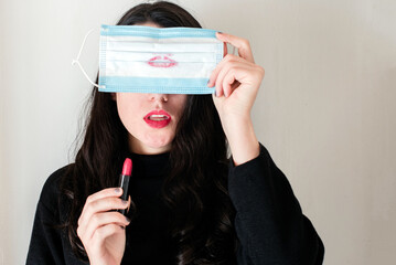 Una mujer joven morena sujeta una mascarilla quirúrgica azul manchada de pintalabios con una mano...