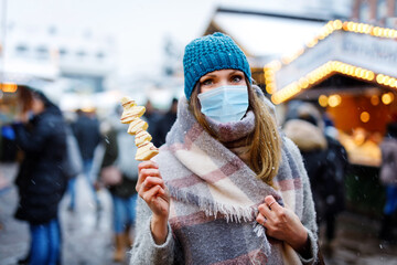 Woman with medical mask eating white chocolate covered fruits on skewer on traditional Christmas market. People with masks as protection against corona virus. Covid pandemic time.
