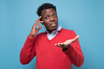 Puzzled african young man having no idea asking what to do. Studio shot on blue wall.