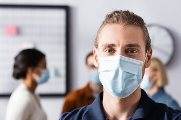 young office manager in medical mask looking at camera near multicultural colleagues on blurred background