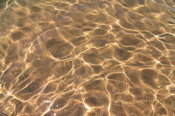 Fototapeta na wymiar Distorted texture of sand under water. Transparent water ripples, sand waves and glare of sunlight. Seabed background
