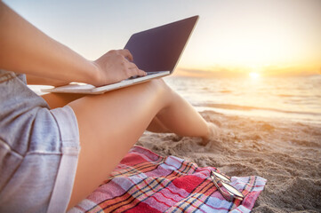 Close-up of a slender young woman on the beach at the sunset time by the sea works on her laptop holding him on her lap. Freelance concept for remote work and work while on vacation