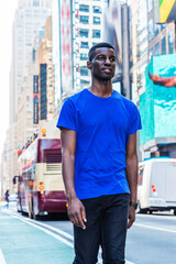 Young African American Man traveling in New York City, wearing blue T shirt, black pants, walking on busy street in Times Square of Manhattan. High buildings, billboards, cars on background..