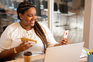 Happy african american woman eating and checking report on smart cell phone in workstation. meal,...