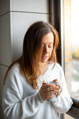 Young woman watches from her home window while holding a hot mug