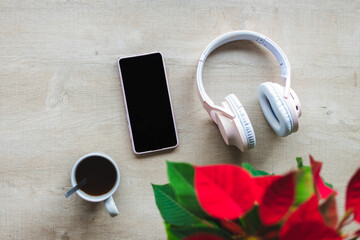 Top view of a wooden table with a poinsettia, wireless headphones, mobile phone and a cup with coffee