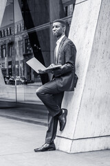 Young African American Businessman working in New York City, wearing suit, white shirt, leather shoes, standing against column on street outside office building, holding laptop computer, thinking..