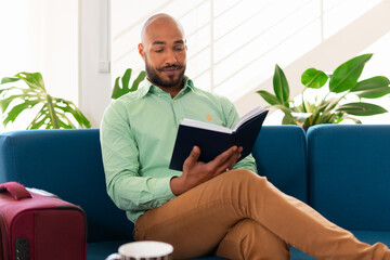 Charming african american man reading a book on sofa of business lounge. Executive, travel, specialist, corporate concept..