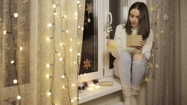 Young woman in knitted sweater and socks wrapping Christmas present in box and sitting in cozy room decorated for winter holidays 