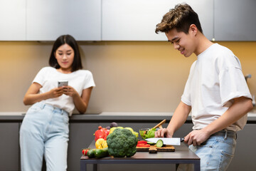 Asian Husband Cooking While Wife Texting On Phone In Kitchen