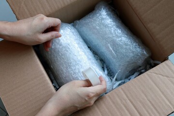 Woman’s hands packing fragile items with plastic air bubble wrapped and adhesive tape and putting...