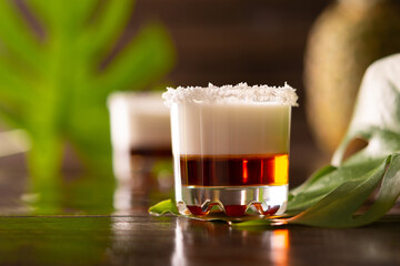 Tropical alcoholic shots decorated with coconut flakes, monstera flower with coconut and pineapple on brown wooden background