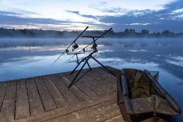 A quiet lake in the fog, two fishing rods on the racks, a wooden pontoon, carpfishing.
