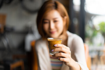 Blurred image of short-haired smiling young Asian woman holding a glass cup of coffee milk in left hand during sitting at a table in a coffee shop. Focus on a hand holding a glass cup of coffee milk.