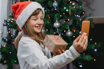 Online congratulations, conferences, chats on holidays. Congratulations from family and friends. A cute smiling girl in a Christmas hat is chatting online by phone with friends.