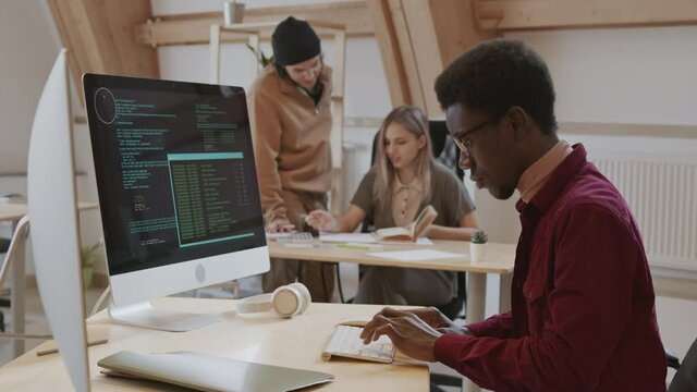 Medium shot of young African male program developer wearing casual clothes sitting at workplace and coding on computer while his colleagues talking