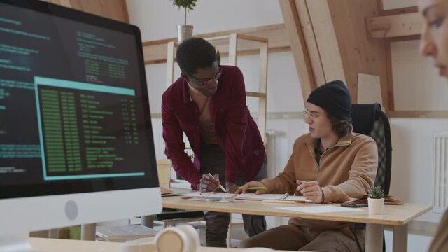 Panning of young Caucasian man and African-American man discussing new design project while young Caucasian female program developer sitting at workplace and coding on computer