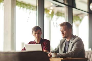 Two business partners, middle aged and young ones, sitting at table in cafe and discussing business project presented on laptop computer over coffee