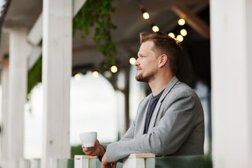 Side view of thoughtful middle aged man leaning on balcony railing at restaurant and drinking coffee