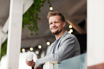 Portrait of handsome middle aged man leaning on balcony railing at restaurant, holding coffee cup and looking at camera