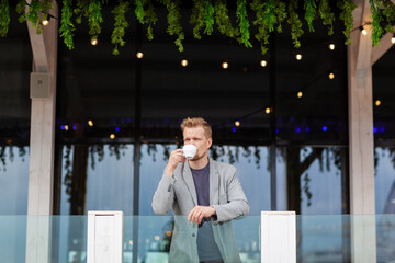 Wide shot of middle aged man leaning on balcony railing, looking away thoughtfully and drinking coffee