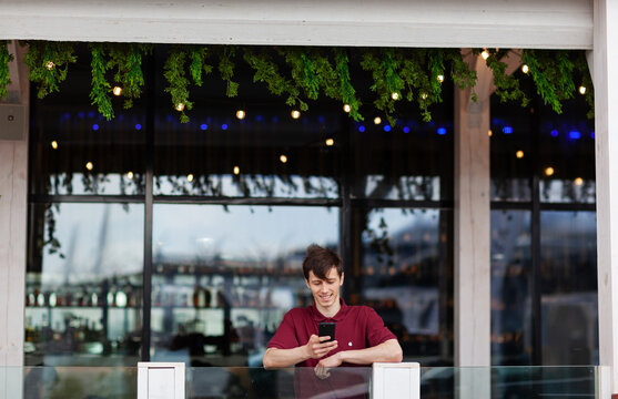 Wide Shot Of Smiling Young Man Leaning On Balcony Railing At Restaurant And Texting On Cell Phone