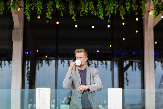 Wide shot of middle aged man leaning on balcony railing, looking away thoughtfully and drinking coffee