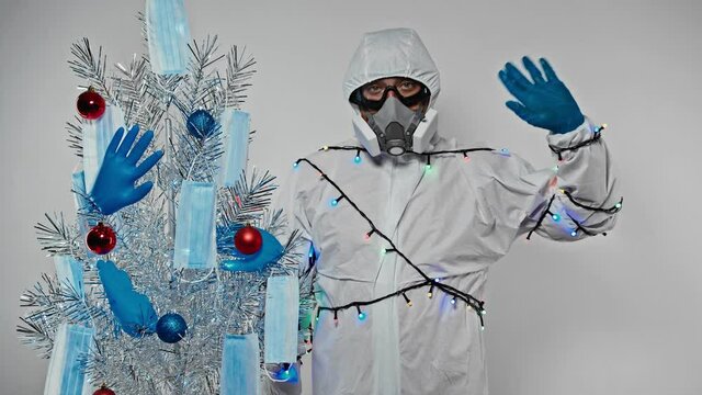 Virusologist (doctor,scientist) wearing a protective suit and garland waves a hand standing next to Christmas tree decoreted with medical masks, gloves and balls.Medical masks on the Christmas tree