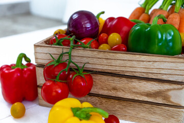 Une caisse de légumes fraiche avec des tomates, des oignons,des poivrons,des carottes,des tomates cerises avec de belles couleurs éclatantes rouges, jaunes,vert et oranges sur un fond de bardage blanc
