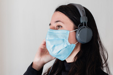 Una mujer joven morena escuchando música con unos auriculares grises mientras lleva una mascarilla...