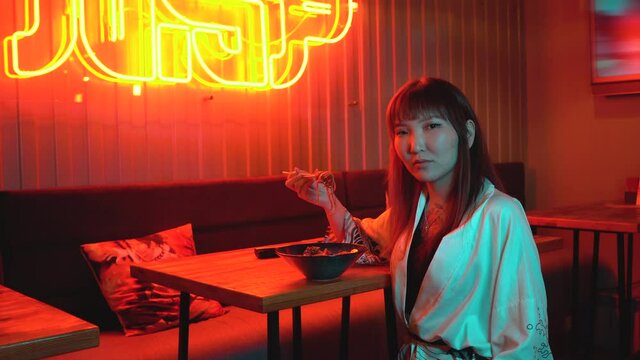 Young fashionable asian woman eating udon noodles with chopsticks at a fast food restaurant. A beautiful girl is sitting in a bar or club against the backdrop of a neon sign. Red neon hieroglyphs.