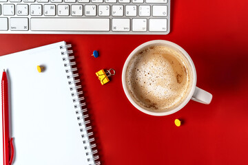 Top view of the office desk. A workspace with an empty notebook, keyboard, stationery, and coffee cup on a red background.
